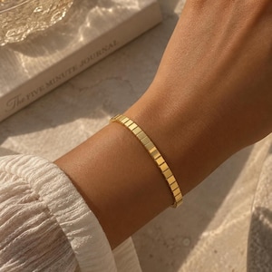 May include: A gold-toned bracelet featuring a series of connected square links that form a flexible, uniform band. The bracelet has a polished, reflective surface and a consistent geometric design that wraps around the wrist in a continuous loop.