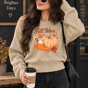 May include: A beige long-sleeve jumper featuring a graphic print with the text Fall Vibes, a large pumpkin, a smaller pumpkin, autumn leaves, and a mug of coffee with whipped cream. The jumper has a wide neckline and a ribbed hem.
