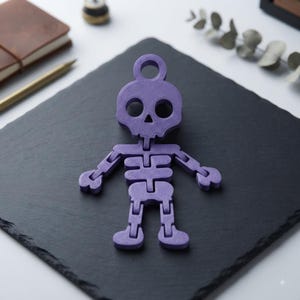 May include: A purple 3D printed articulated skeleton figure with a circular loop at the top of the skull. The skeleton features a stylized head with hollow eye sockets, a triangular nose, and jointed limbs that allow for movement and posing.