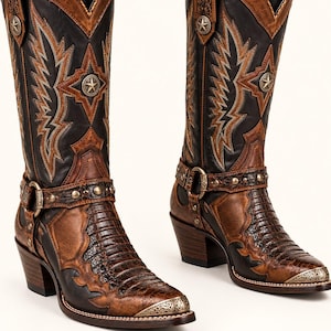Può includere: Un paio di stivali western al ginocchio che combinano pelle nera e marrone vissuta. Gli stivali presentano una tomaia con texture effetto coccodrillo, punte metalliche, emblemi a stella e un cinturino con borchie color ottone.