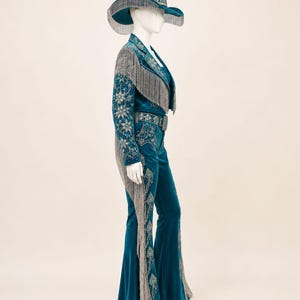 May include: A teal velvet outfit consisting of a cowboy hat, a long-sleeved jacket, and flared trousers. The jacket and hat feature silver fringe, intricate silver embroidery, and clear crystal embellishments. The trousers have matching embroidery.