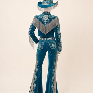 May include: A teal velvet outfit consisting of a cropped jacket and flared pants, both adorned with silver fringe, intricate floral embroidery, and turquoise-colored accents. A matching teal cowboy hat with silver fringe and turquoise details.