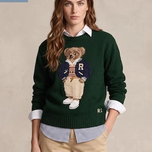 Peut inclure: Un pull en tricot vert foncé à manches longues avec un motif central en intarsia représentant un ours en peluche portant une veste bleu marine avec une lettre R, un gilet à motifs, un pantalon beige et des chaussures blanches, brodé RL en or.