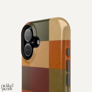 Peut inclure: Une coque de smartphone avec un motif &agrave; carreaux multicolores dans des tons vert olive, beige, orange br&ucirc;l&eacute; et bordeaux fonc&eacute;. La coque pr&eacute;sente un bloc cam&eacute;ra noir avec deux objectifs circulaires, un flash et trois boutons lat&eacute;raux gris.
