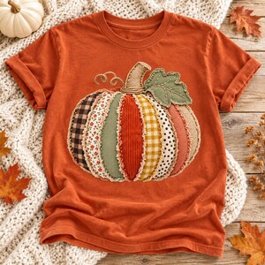 May include: A burnt orange short-sleeve t-shirt featuring a large, central pumpkin applique. The pumpkin is constructed from various fabric patches including plaid, floral, corduroy, gingham, polka dot, and burlap textures with visible stitching.