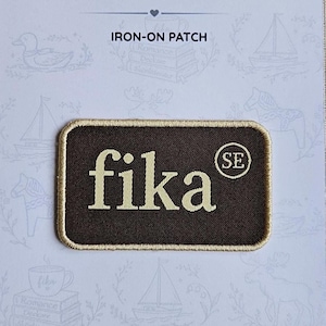 Könnte beinhalten: Ein rechteckiger Aufbügler mit dunkelbraunem Hintergrund und hellbeigem Rand. Der Text fika ist mittig in Hellbeige gestickt, mit den Buchstaben SE in einem Kreis auf der rechten Seite. Der Aufnäher befindet sich auf einer weißen Trägerkarte.
