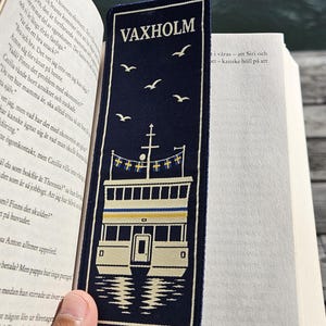 May include: A rectangular dark blue fabric bookmark featuring the white embroidered text VAXHOLM at the top. Below the text is a white and yellow graphic of a ferry boat with small Swedish flags and three white birds flying in the sky above.