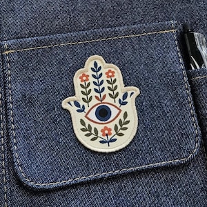 Könnte beinhalten: Ein cremefarbener Hamsa-Hand-Aufnäher mit einem zentralen blauen Augenmotiv, umgeben von orangefarbenen Blumen und grünen Blattzweigen. Der Aufnäher ist auf einer dunkelblauen Jeansstofftasche mit sichtbaren orangefarbenen Nähten befestigt.