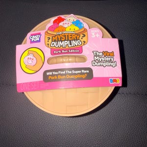 May include: A round, tan, basket-weave textured container holding a mystery toy. The pink packaging features the text "The Original Mystery Dumpling," "Pork Bun Edition," "The Viral Mystery Dumpling!," and "Will You Find The Super Rare Pork Bun Dumpling?"