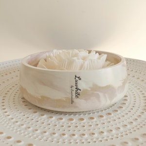 Pode incluir: Uma taça redonda e rasa com acabamento marmoreado em bege e branco contendo uma vela branca em forma de flor. Um rótulo vertical na lateral da taça apresenta o texto Luuwhite by Luxtcandles em fonte cursiva preta sobre a superfície clara.