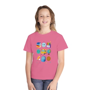 Op de afbeelding: Een roze t-shirt met korte mouwen en een grafische print van schoolthema-iconen, waaronder een stapel boeken, een smiley, een doos kleurpotloden, een gele schoolbus, een wereldbol, een potlood, een blauwe rugzak en een koekje met sterretjes.