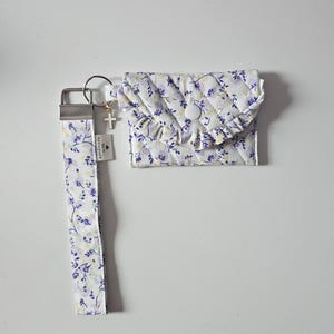 Könnte beinhalten: Ein gestepptes Stofftäschchen mit weißem, violettem und gelbem Blumenmuster, ausgestattet mit einer gerüschten Klappe und einem weißen Druckknopf. Daran befestigt ist ein passendes Stoffarmband mit silbernem Metallverschluss, Kreuzanhänger und Etikett handmade with.