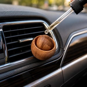 Pode incluir: Um pequeno difusor de aroma para carro em madeira, de forma redonda, fixado numa grelha de ventilação. Um conta-gotas de vidro transparente com tampa preta está posicionado sobre a esfera de madeira, libertando uma gota de líquido na superfície.