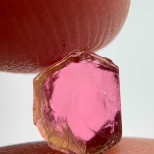 Peut inclure: Une petite pierre précieuse brute de forme hexagonale avec un centre rose vif qui passe à une teinte jaune pâle sur les bords. La pierre présente une texture cristalline translucide avec des facettes naturelles et des inclusions.