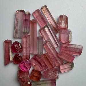 Puede incluir: Una colección de cristales de turmalina en bruto en varios tonos de rosa y rojo. Las piedras presentan formas prismáticas alargadas con estrías verticales y algunos fragmentos irregulares y redondeados, mostrando degradados de color translúcidos.