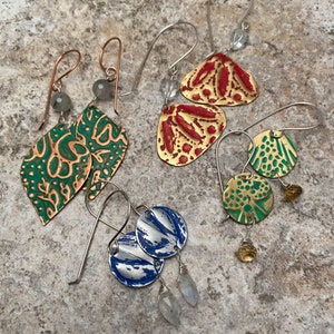 May include: Four pairs of handmade earrings with different patterns and colors. The earrings are made of metal and have a variety of shapes, including leaves, circles, and abstract designs. The earrings are decorated with green, red, blue, and gold paint.