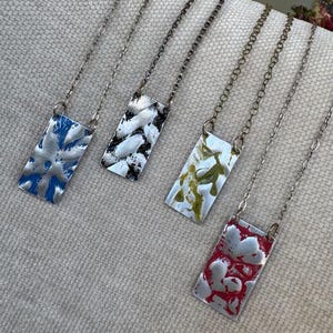 May include: Four rectangular pendant necklaces with textured metal surfaces and chain links. The pendants feature blue, black and white, green and yellow, and red and white designs. The necklaces are displayed on a light gray textured surface.