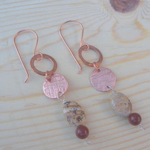 Op de afbeelding: Een paar koperen oorhangers met een gehamerde koperen schijf en een bruine stenen kraal. De oorringen hebben een eenvoudig draadlusontwerp.