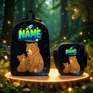 May include: A black backpack and a matching lunch bag, both featuring a space-themed background with stars, a small rocket, and a cartoon illustration of a large brown bear with a cub. The word NAME is printed in large, colorful, bubbly letters.