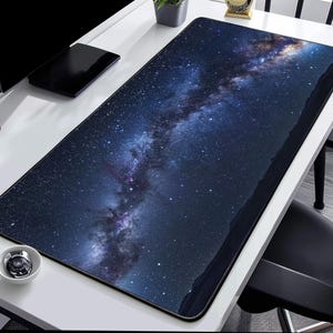 Könnte beinhalten: Eine rechteckige Schreibtischunterlage mit einem hochauflösenden Druck des Nachthimmels, der ein dichtes Sternenfeld und eine leuchtende, wirbelnde Galaxie in tiefen Blau-, Lila- und Weißtönen zeigt, mit glatter Oberfläche und schwarzen Kanten.