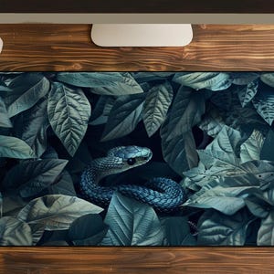 Pode incluir: Um tapete de secretária retangular com uma impressão gráfica detalhada de uma cobra azul-escura enrolada no meio de um grupo denso de folhas verdes e azul-esverdeadas sobrepostas. O tapete tem cantos arredondados e uma borda preta.