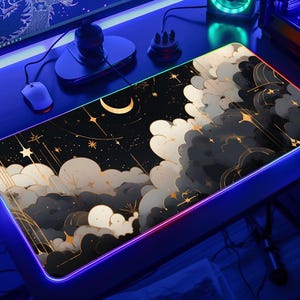 Puede incluir: Una alfombrilla de escritorio rectangular con fondo negro y un diseño celestial de nubes blancas y grises, una luna creciente y estrellas doradas. La alfombrilla está bordeada por una tira de luz LED que emite una luz azul y violeta vibrante.