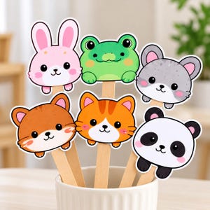 May include: A set of six animal-themed paper cutouts attached to wooden sticks, featuring a pink rabbit, a green frog, a gray cat, a brown cat, an orange tabby cat, and a panda, all arranged in a white ceramic cup.