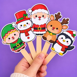 May include: Five holiday-themed paper cutouts attached to wooden sticks, featuring a green-clad elf, a snowman with a red hat, Santa Claus, a brown reindeer with antlers, and a penguin wearing a red Santa hat, all with colorful cartoon designs.