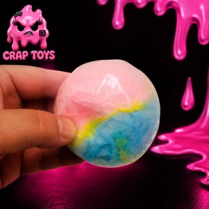 May include: A round, translucent stress ball featuring a marbled blend of pink, yellow, and blue colours. The surface has a textured, cracked appearance. The logo CRAP TOYS with a pink, angry, dripping character icon is visible in the upper left corner.