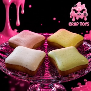 May include: Four square-shaped pastries with smooth, colored glazes in light pink, yellow, white, and pale green, each topped with tiny dark specks. They are arranged on a clear, textured glass pedestal stand against a black background with pink slime.