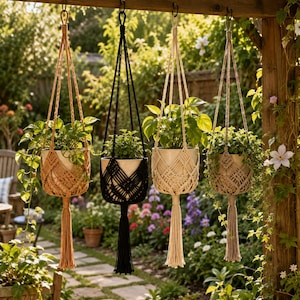 May include: Four macrame plant hangers in tan, black, cream, and beige, each holding a potted green plant. The hangers feature intricate knotted patterns and long tassels at the bottom, suspended from a wooden beam in an outdoor garden setting.