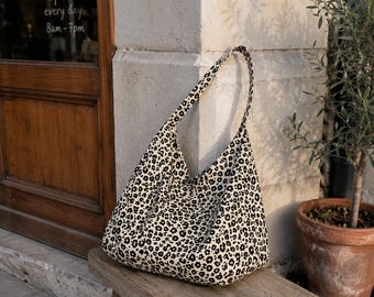 Puede incluir: Un bolso de hombro estilo hobo hecho de tela color hueso con un patrón floral negro repetitivo. El bolso tiene una sola correa integrada y una silueta holgada y sin estructura que se estrecha hacia la abertura superior.
