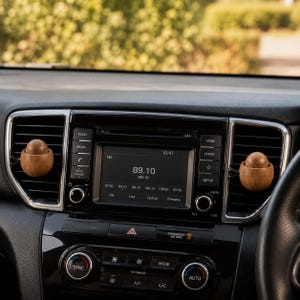 Puede incluir: Dos clips ambientadores de coche de madera de color marrón claro y forma redonda están sujetos a las lamas horizontales de las rejillas de ventilación del salpicadero. La consola central muestra la hora 10:47 y la frecuencia 89.10.