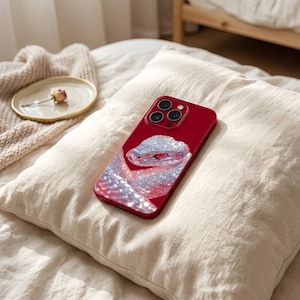 Op de afbeelding: Een dieprode smartphonehoes met een opvallende, sprankelende, zilverkleurige slangafbeelding met een rood oog. De hoes heeft een uitsparing voor een camera met drie lenzen en een gladde, glanzende afwerking die het licht reflecteert.