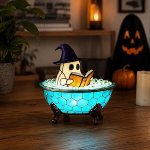 May include: A stained glass lamp shaped like a bathtub with a blue honeycomb pattern, featuring a white ghost figure wearing a purple witch hat and reading an orange book. The bathtub is filled with clear glass beads and stands on four dark metal feet.