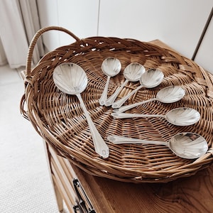May include: A set of seven silver-toned spoons with scalloped bowl designs and decorative handles. The spoons vary in size, with one large serving spoon and six smaller spoons, all featuring a polished metallic finish and intricate detailing.