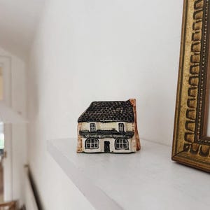 May include: A miniature ceramic building figurine featuring a dark brown textured roof and cream-coloured walls. The front facade displays two ground-floor windows, a central door, and the text "VILLAGE STORES" painted in black above the storefront.