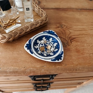 Puede incluir: Un plato triangular de cerámica con base blanca y motivos florales azules pintados a mano. El plato contiene varios dijes pequeños de color dorado con forma de estrella y cuentas blancas similares a perlas en el centro.