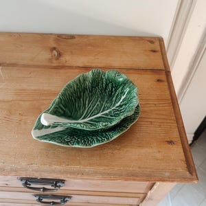 Puede incluir: Un plato de cerámica con forma de hoja de col verde y tallo blanco. El plato presenta una superficie texturizada y veteada que imita la apariencia de una hoja real, con un acabado brillante y un color verde intenso con matices.