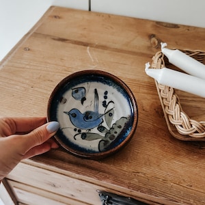 May include: A small, round ceramic dish with a dark brown rim and a cream-coloured centre. The interior features a hand-painted blue bird perched on a branch, accompanied by a small blue butterfly and dark green foliage accents against a blue border.