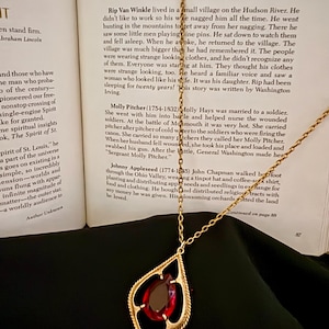 May include: A gold-toned necklace featuring a teardrop-shaped pendant with a twisted metal frame and a central deep red gemstone. The pendant hangs from a delicate gold-toned chain, positioned against an open book titled America's Folk Heros.