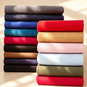 Op de afbeelding: Twee stapels gevouwen stoffen in diverse effen kleuren, waaronder antraciet, marineblauw, koningsblauw, teal, rood, zwart, beige, lichtroze, lichtblauw en olijfgroen. De textielstukken hebben een zachte, matte en licht pluizige textuur.