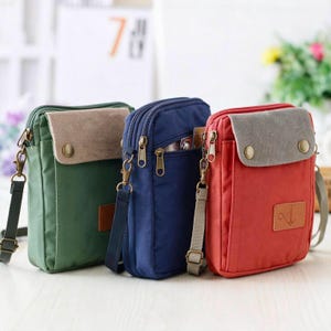 Puede incluir: Tres bolsos bandolera rectangulares pequeños en verde, azul marino y rojo. Cada bolso cuenta con una solapa superior con dos botones a presión color latón, un compartimento principal con cremallera y un parche con un ancla.