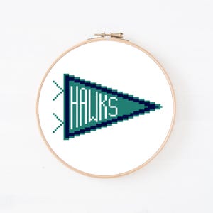 May include: A circular wooden embroidery hoop containing a cross-stitch design of a teal and dark blue triangular pennant flag. The word HAWKS is stitched in white pixelated letters inside the pennant, which features two small decorative tails.