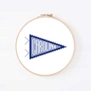 Op de afbeelding: Een ronde houten borduurring met witte stof en een pixel art kruissteekontwerp. Het ontwerp toont een lichtblauwe driehoekige wimpel met een donkerblauwe rand en het woord CAROLINA in witte blokletters.