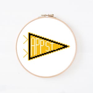 May include: A circular wooden embroidery hoop containing a white fabric panel with a pixel art design of a yellow triangular pennant. The pennant features a thick black border and the white text APPST written in a blocky, pixelated font style.