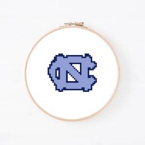 Op de afbeelding: Een ronde houten borduurring die een witte stoffen achtergrond omlijst. Gecentreerd op de stof staat een logo in pixel-art-stijl met de letters NC in lichtblauw met een donkerblauwe omlijning, wat een blokkerige, digitale esthetiek creëert.