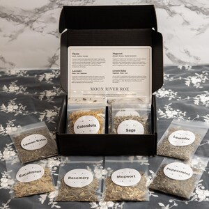 May include: A black cardboard box containing dried herbs in clear plastic bags, labelled Calendula, Sage, Lemon Balm, Eucalyptus, Rosemary, Mugwort, and Peppermint. An informational card titled Moon River Roe is inside the box lid.