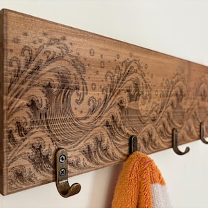 May include: A rectangular wooden wall rack featuring a dark wood-burned design of rolling ocean waves and bubbles. The board is mounted with four bronze-colored metal hooks, one of which holds a folded orange and white textured fabric towel.