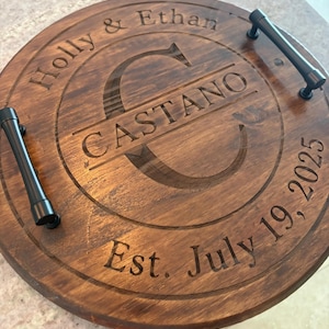 May include: A round, dark brown wooden serving tray with two black metal handles. The surface is engraved with the names Holly & Ethan, the surname CASTANO centered over a large letter C, and the text Est. July 19, 2025 along the bottom edge.